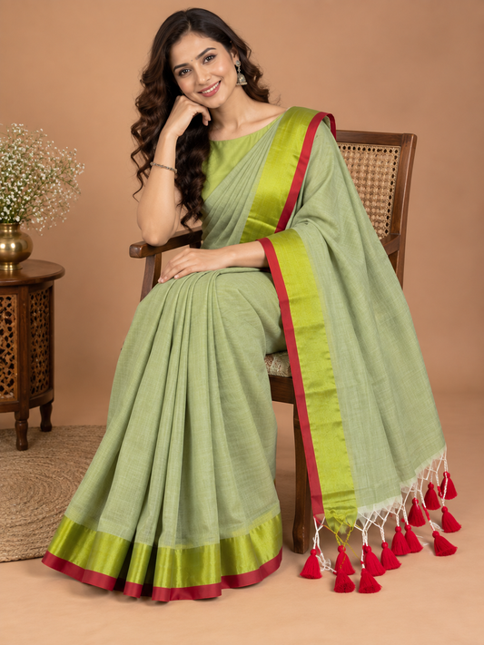 A woman seated on a wooden chair wearing a light olive green saree with a bright yellow-green border and red tassel accents, posed against a warm beige studio backdrop.