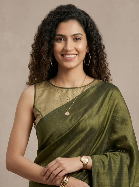 A woman with curly dark hair wearing a draped olive green silk saree with a sheer mesh sleeveless blouse, posed against a neutral beige background.
