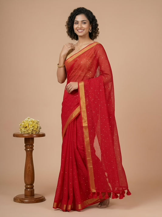 A woman modeling a red saree with gold border and small gold motifs, standing against a beige studio background next to a small wooden pedestal with yellow flowers.