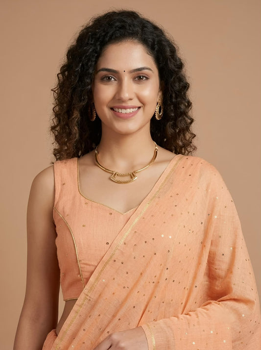 A woman with curly dark hair wearing a peach-colored traditional Indian sari with gold sequins and a matching sleeveless blouse, posed against a solid warm beige background.