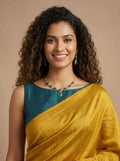 A woman with curly dark hair wearing a mustard yellow and teal draped sari with a matching gold necklace and earrings, posed against a solid beige background.