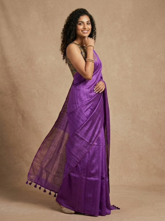 A woman modeling a purple silk saree with a matching sleeveless blouse, standing against a textured beige backdrop.