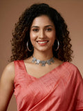 A smiling woman with curly dark hair wearing a coral pink sari and a silver-toned statement necklace, photographed against a warm brown studio background.