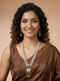 A smiling woman with curly dark hair wears a brown sari and a long layered silver-toned necklace with a decorative pendant, along with matching drop earrings, against a plain beige background.