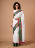 A woman modeling a white saree with a green and magenta border, paired with a white sleeveless blouse, set against a solid beige studio background.