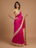 A woman modeling a bright pink silk saree with a gold zari border and a matching gold blouse, set against a solid warm brown studio background.