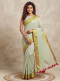 A woman modeling a pale sage green saree with a wide gold zari border and red tassel accents, posed against a neutral beige studio backdrop.