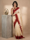 A woman wearing a cream-colored silk saree with a deep red tie-dye border and pallu, standing next to a gray concrete pedestal with a gold sculpture against a beige background.