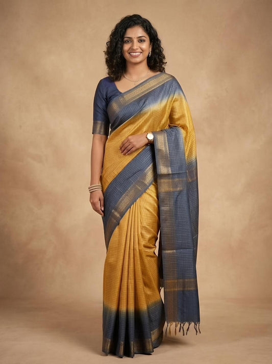 A woman modeling a mustard yellow and navy blue saree with a matching navy blue blouse, standing against a warm beige studio backdrop.