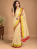 A woman models a beige saree with a wide yellow and red border, standing against a solid beige studio background with a small wooden side table and flowers on the left.