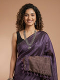 A woman with curly dark hair models a purple silk saree with a textured brown pallu and a black sleeveless blouse, accessorized with silver jewelry, against a solid beige background.