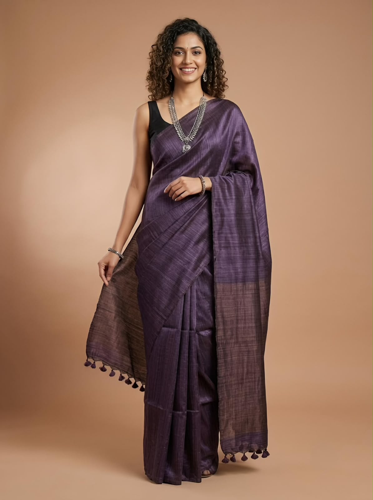 A woman modeling a purple silk saree with a black sleeveless blouse, standing against a solid warm beige studio background.