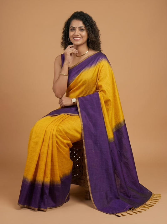 A woman seated on a dark stool wearing a yellow and purple tie-dye saree with gold zari detailing, posed against a solid warm beige studio backdrop.