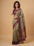 A woman modeling a traditional Indian saree in a studio setting. The saree has a muted sage green body with purple borders, gold zari motifs, and a matching purple blouse.