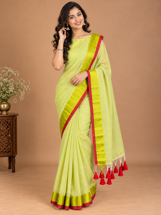 A woman models a light lime green saree with a red border and tassel trim, standing against a solid beige studio background.