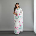 A woman modeling a white saree with a pink and green floral print, standing against a plain gray studio background.