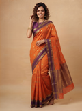 A woman modeling an orange silk saree with purple borders and gold motifs, standing against a solid beige studio backdrop.