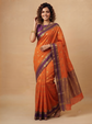 A woman modeling an orange silk saree with purple borders and gold motifs, standing against a solid beige studio backdrop.