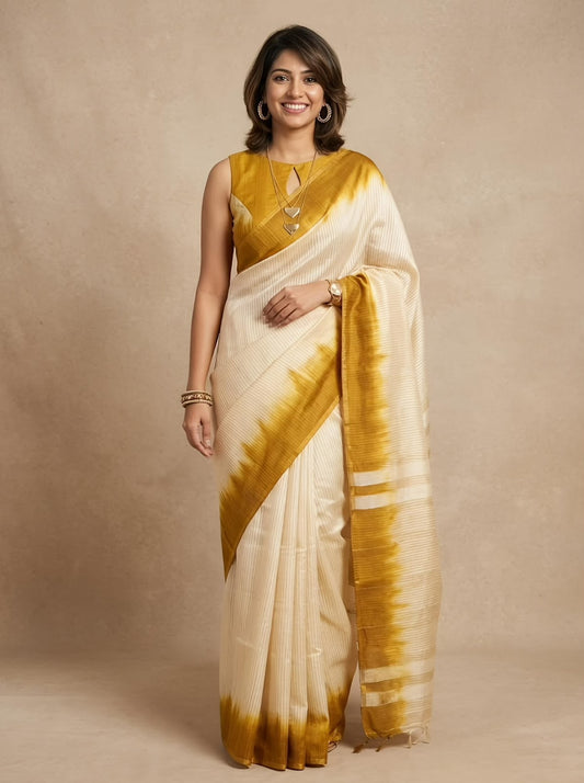 A woman modeling a cream-colored silk saree with a mustard yellow tie-dye border and a matching yellow blouse, standing against a neutral beige studio backdrop.