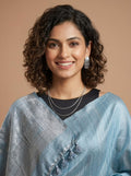 A woman with curly dark hair smiling, wearing a light blue textured sari with tassel details over a black top, set against a solid warm beige background.
