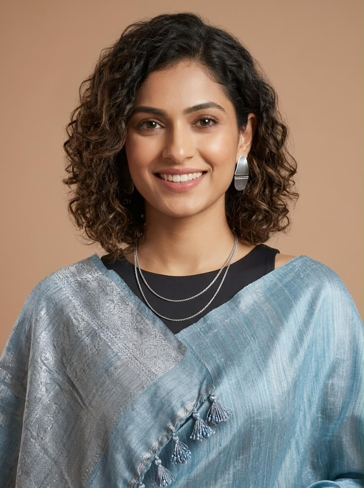 A woman with curly dark hair smiling, wearing a light blue textured sari with tassel details over a black top, set against a solid warm beige background.