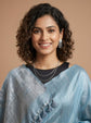 A woman with curly dark hair smiling, wearing a light blue textured sari with tassel details over a black top, set against a solid warm beige background.