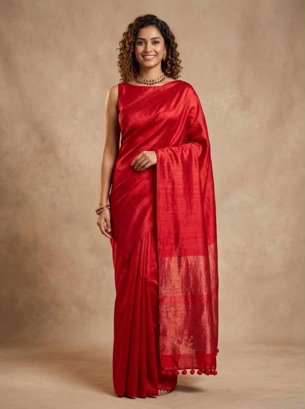 A woman modeling a vibrant red silk saree with a matching red sleeveless blouse, standing against a textured beige studio backdrop.