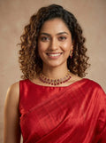 A smiling woman with curly brown hair wearing a red sari and a matching red and gold necklace, photographed against a warm beige studio background.