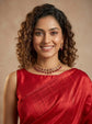 A smiling woman with curly brown hair wearing a red sari and a matching red and gold necklace, photographed against a warm beige studio background.