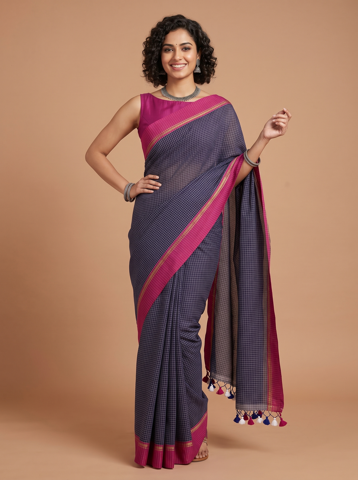 A woman modeling a dark purple and magenta checkered saree with gold borders and tassel accents, posed against a solid beige studio background.