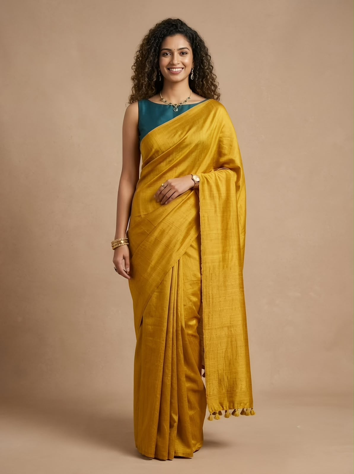 A woman modeling a mustard yellow silk saree with a teal blue sleeveless blouse, standing against a solid beige studio backdrop.