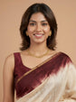 A woman wearing a maroon and beige tie-dye silk saree with a gold border, styled with a gold necklace and earrings, against a solid beige background.