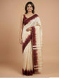 A woman modeling a cream-colored silk saree with a deep maroon tie-dye border and matching blouse, set against a solid beige studio background.