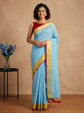 A woman modeling a light blue silk saree with a gold and maroon border, paired with a maroon sleeveless blouse, standing against a warm beige studio backdrop.