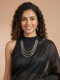 A smiling woman with curly dark hair models a multi-strand silver chain necklace with triangular pendant details over a black sleeveless top and a black draped fabric. The background is a solid warm beige studio backdrop.