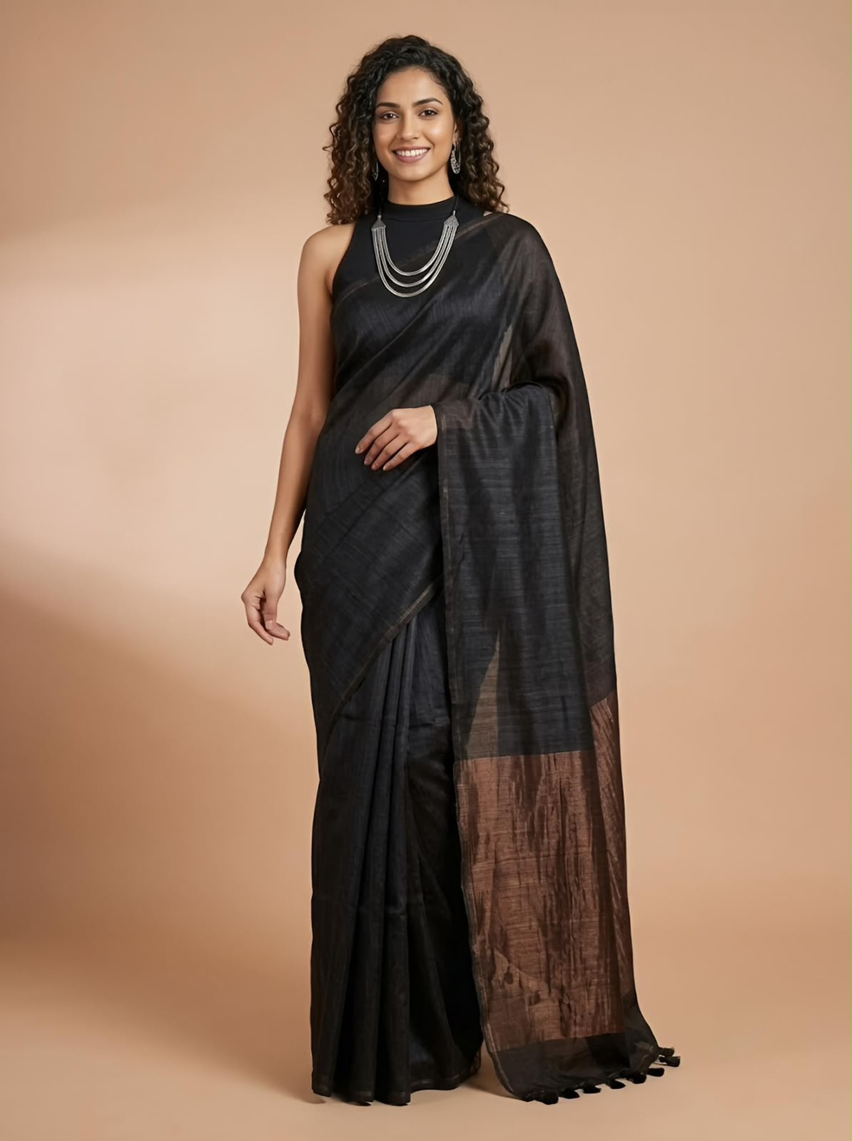 A woman modeling a black silk saree with a brown border, standing against a solid beige studio background.