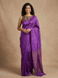 A woman modeling a purple silk saree with a matching embroidered blouse, standing against a textured beige studio backdrop.