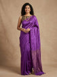 A woman modeling a purple silk saree with a matching embroidered blouse, standing against a textured beige studio backdrop.