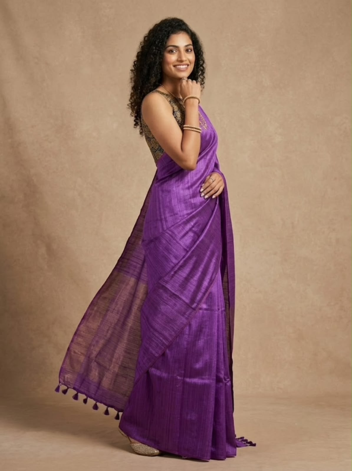 A woman modeling a purple silk saree with a matching sleeveless blouse, standing against a textured beige backdrop.