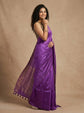 A woman modeling a purple silk saree with a matching sleeveless blouse, standing against a textured beige backdrop.