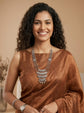 A smiling woman with curly dark hair wearing a bronze-brown sleeveless top and a layered silver statement necklace, photographed against a warm beige studio background.