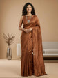 A woman modeling a rust-brown silk saree with a matching sleeveless blouse, standing in a warm beige studio setting with a sofa and vase in the background.