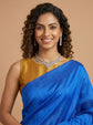 A woman with curly dark hair wearing a royal blue sari with a gold blouse, styled with a silver necklace and earrings, set against a solid beige background.