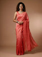 A woman modeling a coral pink silk saree with a subtle checkered pattern and tassel border, set against a solid warm brown studio background.