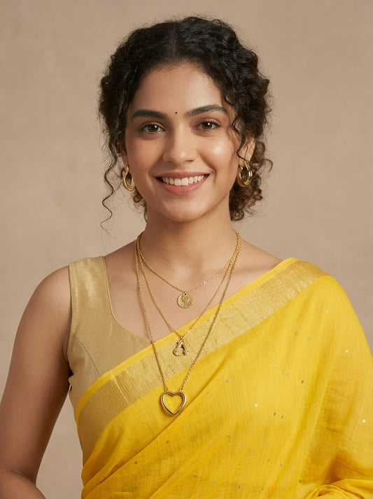 A woman with curly dark hair wearing a bright yellow saree with a gold blouse and gold jewelry, including layered necklaces and earrings, against a solid beige background.