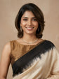 A woman wearing a beige and black tie-dye silk saree with a gold border, styled with a gold necklace and earrings, against a warm beige background.
