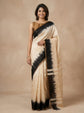 A woman modeling a beige and black tie-dye saree with a solid beige blouse, standing against a textured beige studio backdrop.