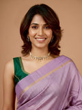 A woman wearing a lavender purple and green saree with gold detailing, styled with a gold necklace and earrings, against a solid beige background.
