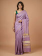 A woman modeling a lavender purple silk saree with gold zari borders and a beige and gold striped pallu, paired with a dark green blouse, against a solid beige studio background.