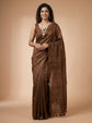 A woman modeling a brown silk saree with a matching sleeveless blouse, standing against a solid beige studio background.